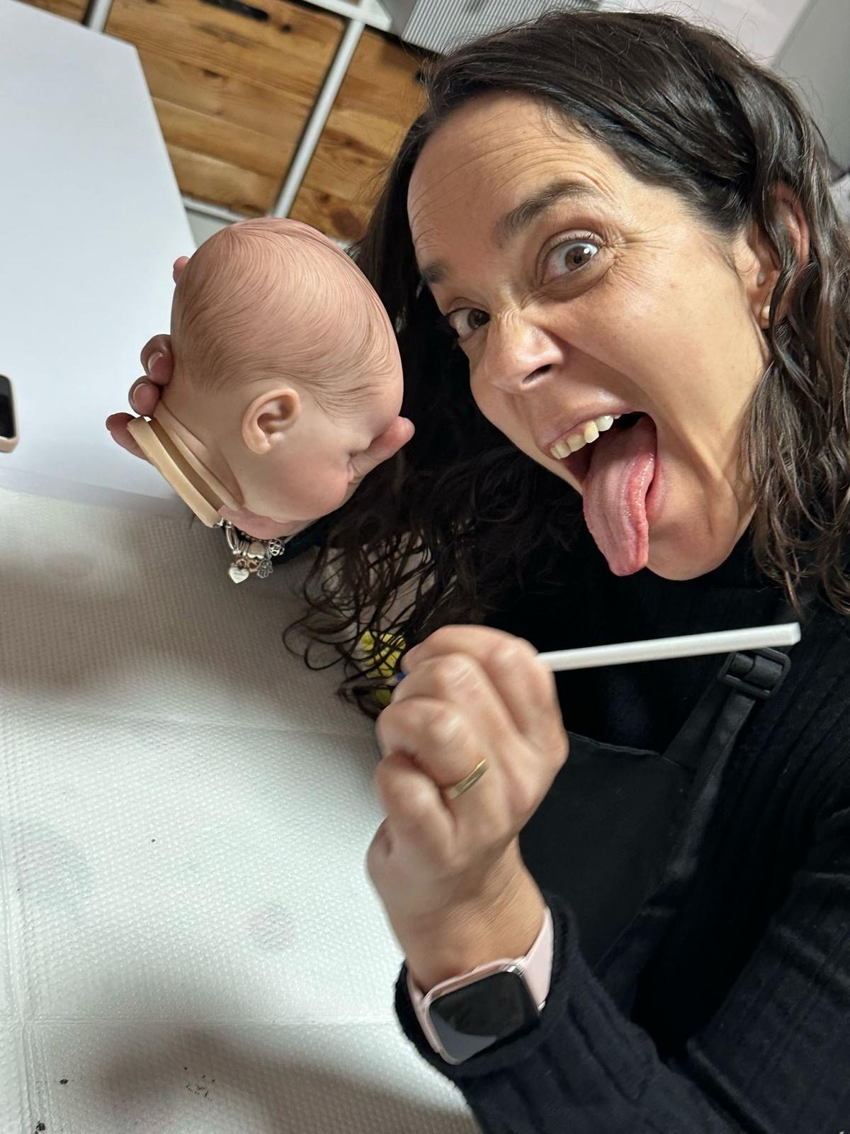 Miriam sacando la lengua en su mesa de trabajo, con un pincel en la mano y una cabeza de reborn a medio pintar
