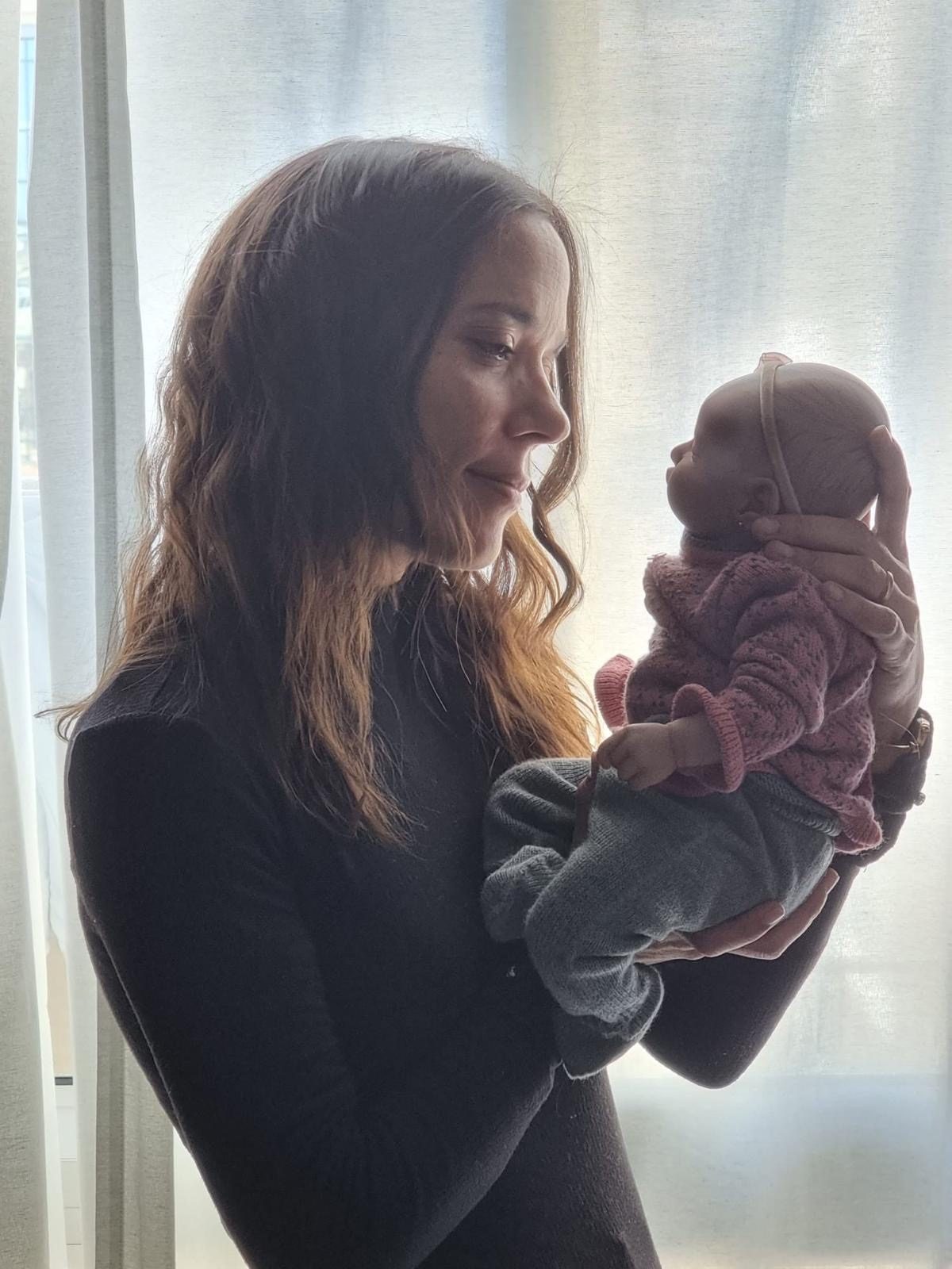 Miriam junto a una ventana, mirando con una sonrisa a una bebé reborn vestida con jersey rosa